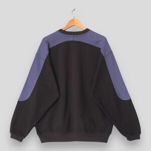 May include: Black sweatshirt with contrasting blue panels on the shoulders and sleeves. The back of the garment is visible, hanging from a wooden hanger. The sweatshirt has a crew neck and a relaxed fit.