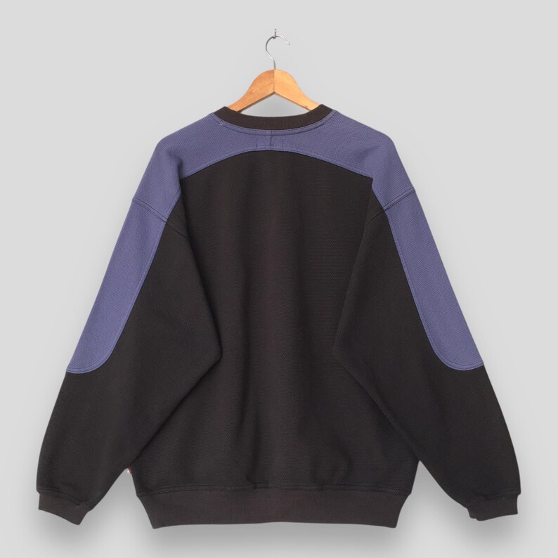 May include: Black sweatshirt with contrasting blue panels on the shoulders and sleeves. The back of the garment is visible, hanging from a wooden hanger. The sweatshirt has a crew neck and a relaxed fit.