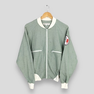 Può includere: Giacca bomber a righe verdi e bianche con colletto, polsini e cerniera bianchi. Presenta una toppa rossa e bianca sulla manica. La giacca è appesa a una gruccia di legno.