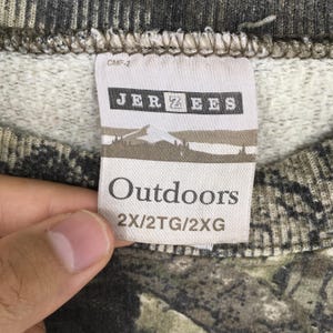 May include: Close-up of a tag from a camouflage-patterned sweatshirt. The tag reads "JERZEES Outdoors" with a mountain and tree graphic. The sweatshirt is a mix of green, brown, and gray.