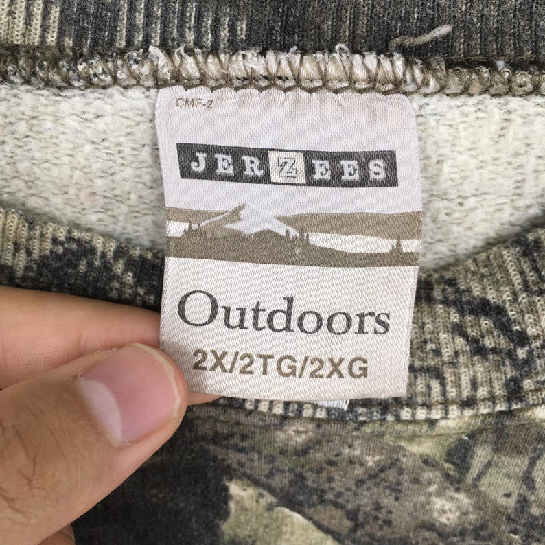 May include: Close-up of a tag from a camouflage-patterned sweatshirt. The tag reads "JERZEES Outdoors" with a mountain and tree graphic. The sweatshirt is a mix of green, brown, and gray.