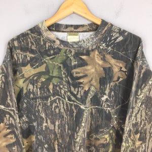 Puede incluir: Sudadera con estampado de camuflaje y cuello redondo, con una mezcla de tonos marrones, verdes y beiges. El dise&ntilde;o incorpora motivos de hojas y ramas, creando un efecto de camuflaje de bosque. La prenda se muestra en una percha de madera.
