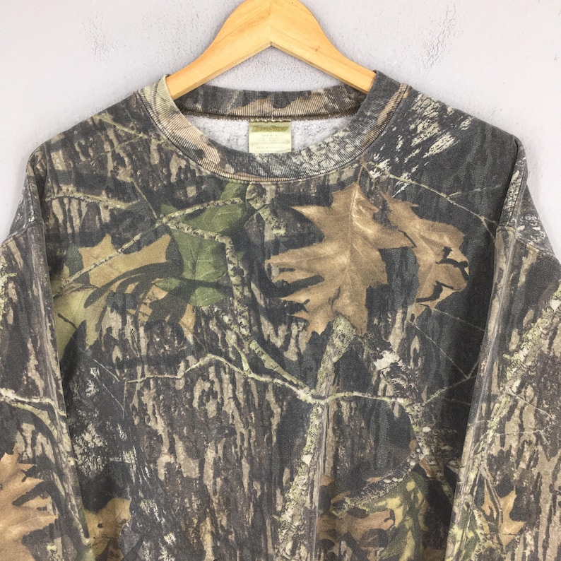 Puede incluir: Sudadera con estampado de camuflaje y cuello redondo, con una mezcla de tonos marrones, verdes y beiges. El dise&ntilde;o incorpora motivos de hojas y ramas, creando un efecto de camuflaje de bosque. La prenda se muestra en una percha de madera.