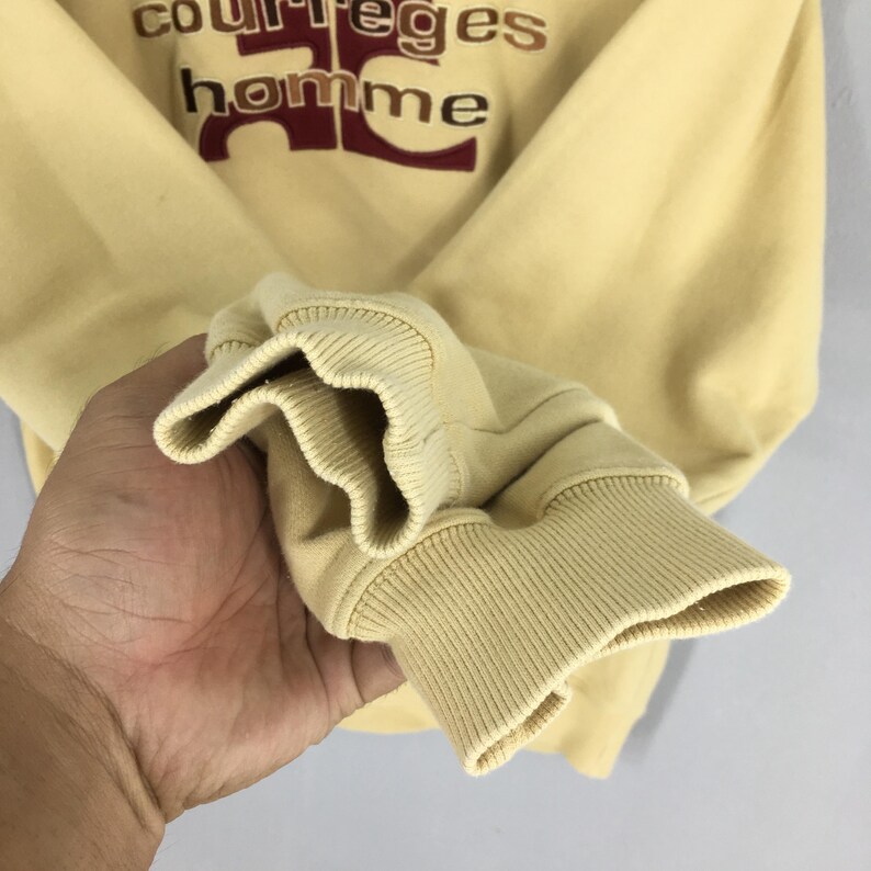 May include: A light yellow sweatshirt with ribbed cuffs and the text "courreges homme" in brown and burgundy. The sweatshirt has a crew neck and long sleeves. The fabric appears to be a soft, knit material.