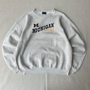 May include: A light gray crewneck sweatshirt featuring the embroidered text MICHIGAN EST. 1817 in navy blue and gold lettering. The garment has long sleeves, a ribbed collar, and a classic athletic fit with a soft, heathered fabric texture throughout.
