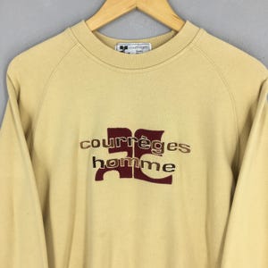 May include: A tan-colored Courr&egrave;ges sweatshirt with the brand name and "homme" in a burgundy and brown embroidered design. The sweatshirt has a crew neck and long sleeves. The Courr&egrave;ges logo is on the collar.