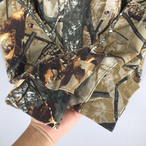 May include: A close-up of a camouflage-patterned hoodie sleeve. The fabric features a mix of brown, tan, and green hues, mimicking a forest environment. The cuff is visible, showing the detailed stitching and the texture of the material.