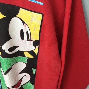 May include: Red sweatshirt featuring a graphic of Mickey Mouse. The design includes Mickey's face and hands in a colorful, rectangular frame. The sweatshirt is a solid red color.