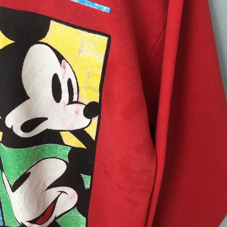 May include: Red sweatshirt featuring a graphic of Mickey Mouse. The design includes Mickey's face and hands in a colorful, rectangular frame. The sweatshirt is a solid red color.