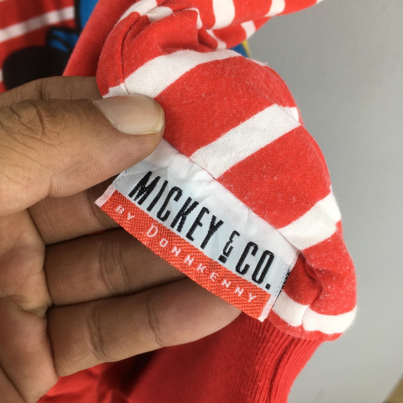 May include: Close-up of a red and white striped fabric with the label "MICKEY & CO. BY DONNKENNY". The fabric appears to be part of a garment, possibly a shirt or sweatshirt, with a ribbed cuff visible.