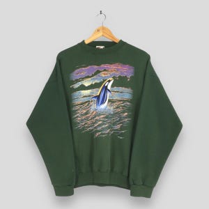 May include: Dark green sweatshirt with a graphic of a breaching orca whale in the ocean under a sunset sky. The orca is black and white with blue accents, and the waves are shades of orange and blue.