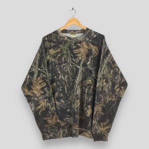 Puede incluir: Una sudadera de camuflaje de manga larga con cuello redondo, colgada de una percha de madera. El dise&ntilde;o presenta tonos de marr&oacute;n, verde y negro, que recuerdan a un entorno forestal. La sudadera est&aacute; hecha de un material suave.