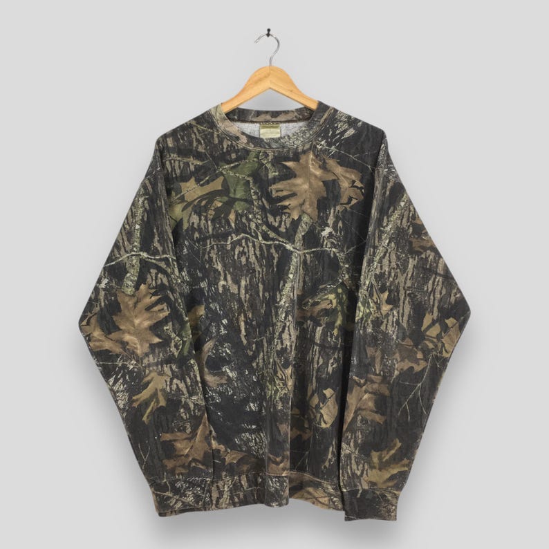Puede incluir: Una sudadera de camuflaje de manga larga con cuello redondo, colgada de una percha de madera. El dise&ntilde;o presenta tonos de marr&oacute;n, verde y negro, que recuerdan a un entorno forestal. La sudadera est&aacute; hecha de un material suave.