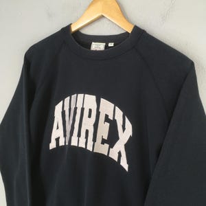 May include: A dark navy blue long-sleeve sweatshirt featuring the word AVIREX printed in large, arched, off-white block letters across the chest. The garment has a crew neckline and is displayed on a wooden hanger against a neutral background.