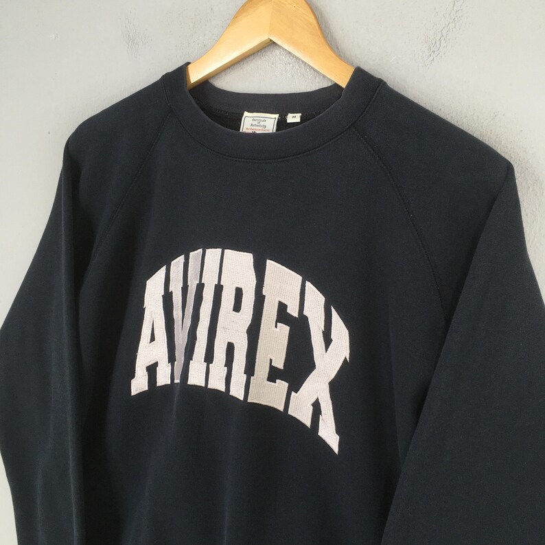 May include: A dark navy blue long-sleeve sweatshirt featuring the word AVIREX printed in large, arched, off-white block letters across the chest. The garment has a crew neckline and is displayed on a wooden hanger against a neutral background.