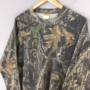 Puede incluir: Una sudadera con estampado de camuflaje en tonos marr&oacute;n, verde y gris, con cuello redondo y mangas largas. El dise&ntilde;o incorpora patrones de hojas y ramas, creando un efecto de camuflaje de bosque. La sudadera est&aacute; colgada en una percha de madera.