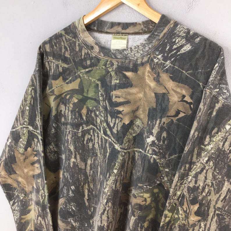 Puede incluir: Una sudadera con estampado de camuflaje en tonos marr&oacute;n, verde y gris, con cuello redondo y mangas largas. El dise&ntilde;o incorpora patrones de hojas y ramas, creando un efecto de camuflaje de bosque. La sudadera est&aacute; colgada en una percha de madera.