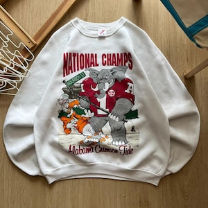 May include: A white crewneck sweatshirt featuring a graphic print of a grey elephant in a crimson jersey labeled with the number 1, standing over an orange duck. The text reads NATIONAL CHAMPS and Alabama Crimson Tide with a Bourbon St sign.