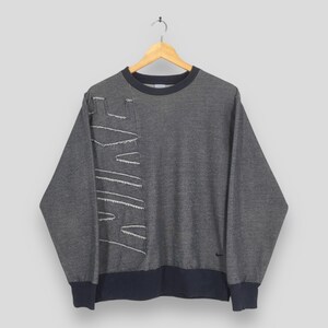 May include: Gray sweatshirt with the Nike logo stitched in white on the left side. The long-sleeved top has a navy blue collar, cuffs, and hem. A small Nike logo is visible on the lower right side.