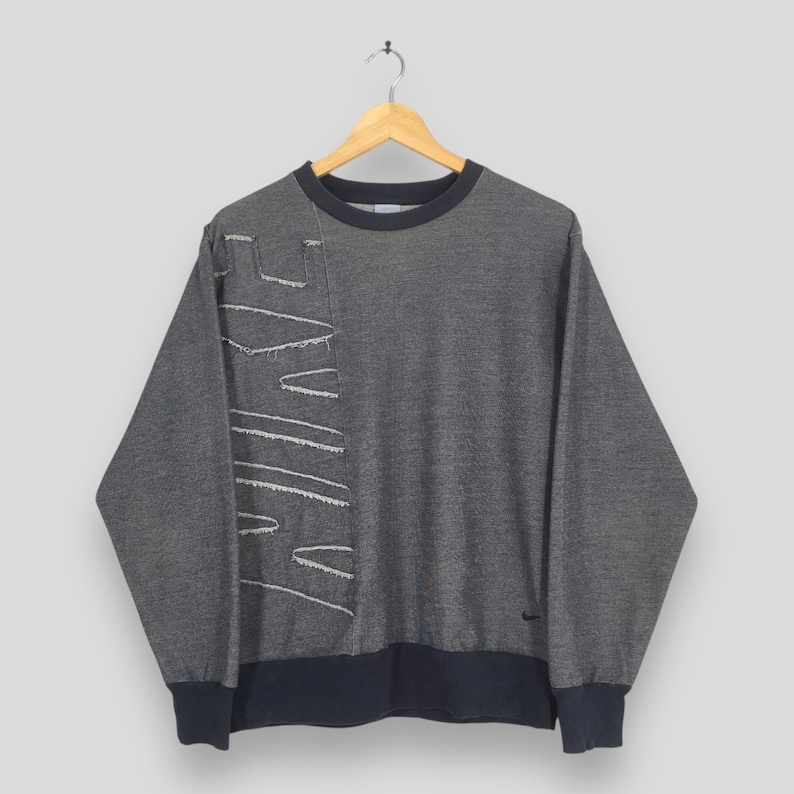 May include: Gray sweatshirt with the Nike logo stitched in white on the left side. The long-sleeved top has a navy blue collar, cuffs, and hem. A small Nike logo is visible on the lower right side.