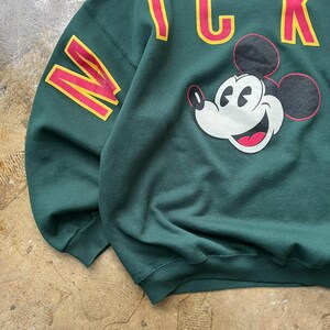 K&ouml;nnte beinhalten: Dunkelgr&uuml;nes Sweatshirt mit Mickey Mouse-Grafik und dem Wort "MICK" in Gelb und Rot. Ein gro&szlig;es "N" in den gleichen Farben befindet sich auf dem &Auml;rmel. Ein Sweatshirt im klassischen Vintage-Stil.