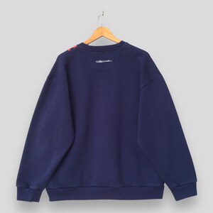 May include: Navy blue sweatshirt with red accents on the shoulder. The back of the sweatshirt is visible, hanging from a wooden hanger. The sweatshirt has a crew neck and a small embroidered design near the neckline. The fabric appears to be a soft, comfortable material.
