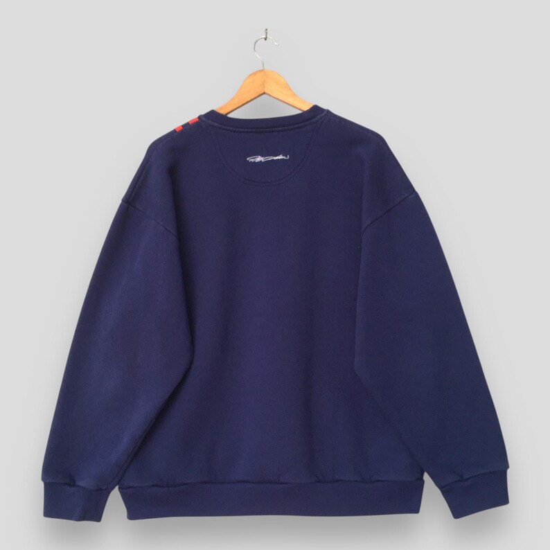 May include: Navy blue sweatshirt with red accents on the shoulder. The back of the sweatshirt is visible, hanging from a wooden hanger. The sweatshirt has a crew neck and a small embroidered design near the neckline. The fabric appears to be a soft, comfortable material.