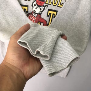 Puede incluir: Una sudadera gris claro de manga larga con pu&ntilde;os acanalados. El pecho presenta un gr&aacute;fico de una mascota lobo con un su&eacute;ter rojo con el texto NCSU, rodeado de letras amarillas que dicen parcialmente WOLF PACK.