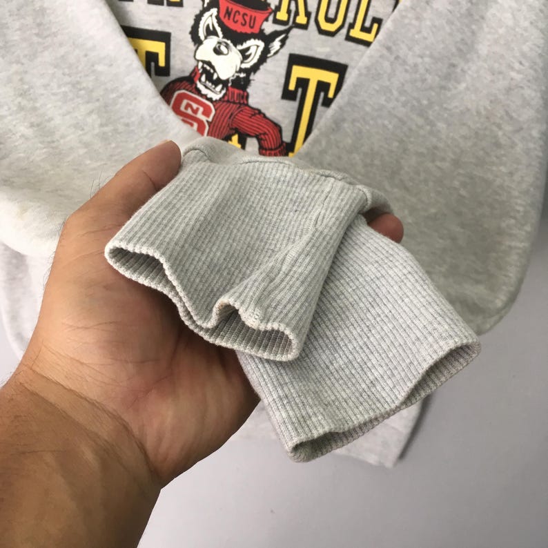 Puede incluir: Una sudadera gris claro de manga larga con pu&ntilde;os acanalados. El pecho presenta un gr&aacute;fico de una mascota lobo con un su&eacute;ter rojo con el texto NCSU, rodeado de letras amarillas que dicen parcialmente WOLF PACK.