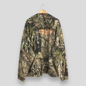 K&ouml;nnte beinhalten: Ein Camouflage-Kapuzenpullover mit einem braunen, gr&uuml;nen und beigen Muster. Die R&uuml;ckseite des Hoodies ist sichtbar und h&auml;ngt an einem Holzb&uuml;gel. Der Stoff hat einen detaillierten Aufdruck von &Auml;sten und Bl&auml;ttern.