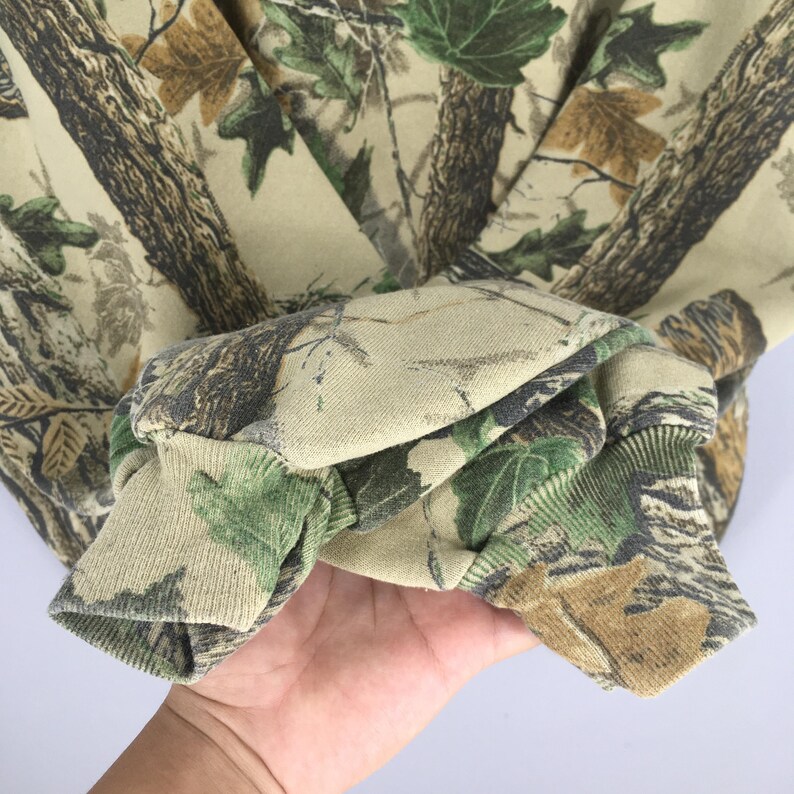 May include: A camouflage-patterned sweatshirt with a forest-like design featuring brown tree trunks, green leaves, and tan backgrounds. The cuffs are folded over, showing the interior fabric. The overall theme is outdoors and hunting.