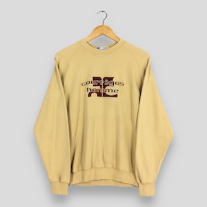 May include: A light tan Courr&egrave;ges homme sweatshirt hanging on a wooden hanger. The sweatshirt features the brand's logo in a burgundy color. The raglan sleeves and crew neck are visible. The fabric appears to be a soft, comfortable material.