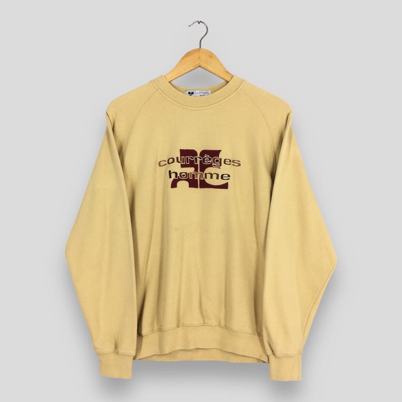 May include: A light tan Courr&egrave;ges homme sweatshirt hanging on a wooden hanger. The sweatshirt features the brand's logo in a burgundy color. The raglan sleeves and crew neck are visible. The fabric appears to be a soft, comfortable material.