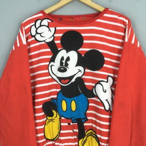 May include: Red and white striped long-sleeve shirt featuring a large graphic of Mickey Mouse. Mickey is depicted in black, white, blue, and yellow. The shirt has red sleeves and is hanging on a wooden hanger.