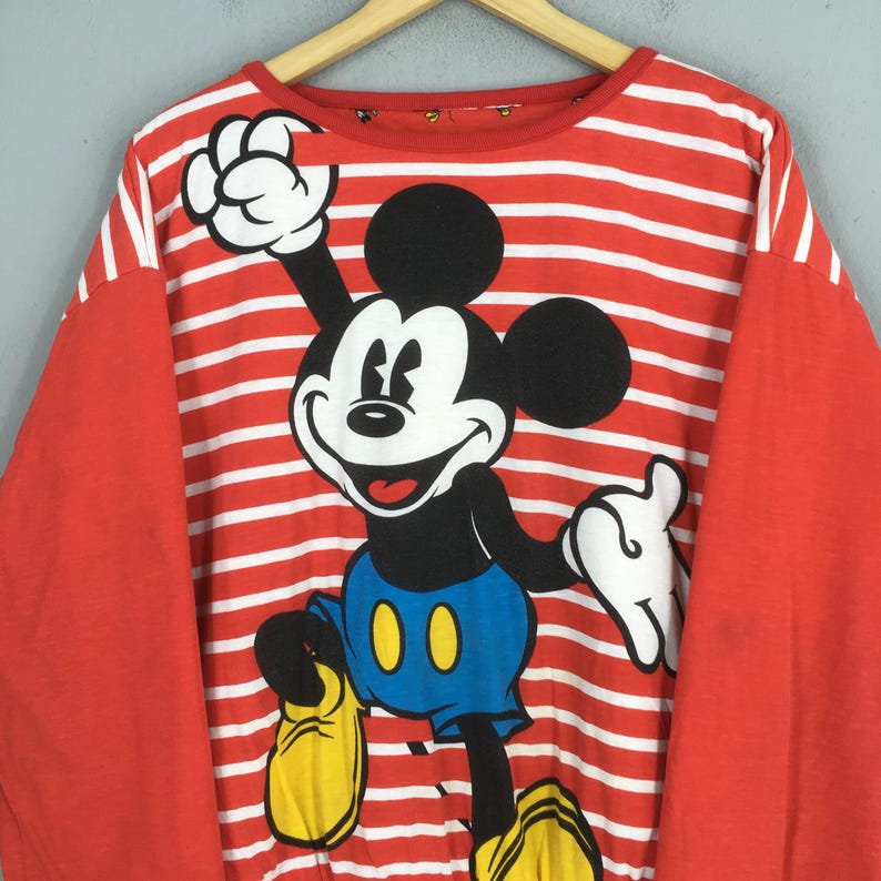 May include: Red and white striped long-sleeve shirt featuring a large graphic of Mickey Mouse. Mickey is depicted in black, white, blue, and yellow. The shirt has red sleeves and is hanging on a wooden hanger.