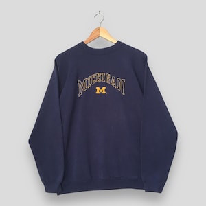 May include: A navy blue crewneck sweatshirt featuring the word MICHIGAN arched in yellow outline lettering above a solid yellow block letter M. The garment has long sleeves with ribbed cuffs and a ribbed hem, displayed on a wooden hanger.