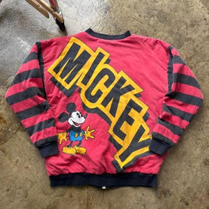 May include: Red and black vintage Mickey Mouse bomber jacket. The jacket features the word "MICKEY" in yellow and black block letters across the back, with a cartoon image of Mickey Mouse. The sleeves have black and red stripes.