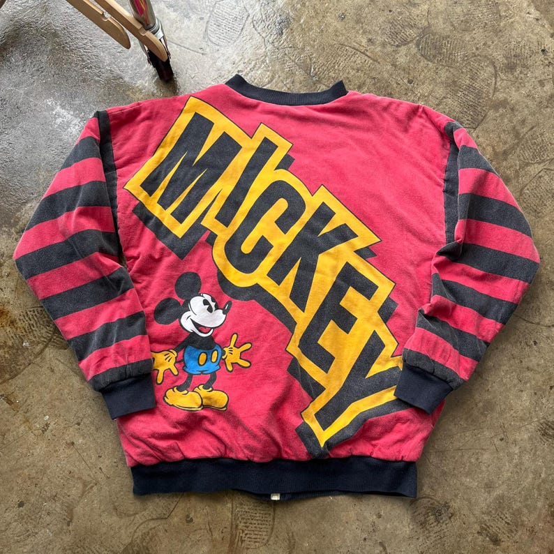 May include: Red and black vintage Mickey Mouse bomber jacket. The jacket features the word "MICKEY" in yellow and black block letters across the back, with a cartoon image of Mickey Mouse. The sleeves have black and red stripes.
