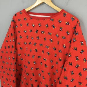 May include: Red long-sleeved sweatshirt with a repeating pattern of cartoon mouse characters in blue, yellow, and black. The sweatshirt has a crew neck with a white inner lining. The garment is hanging on a wooden hanger.