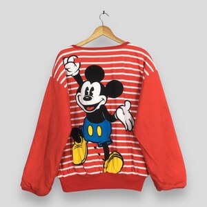 May include: Red and white striped sweatshirt featuring a large Mickey Mouse graphic on the back. The long-sleeved top has red sleeves and a crew neck. The Mickey Mouse graphic is in black, white, yellow, and blue.