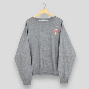 May include: A heather gray sweatshirt with a small embroidered design on the upper left chest. The design appears to be a pink ice cream cone with a brown waffle cone and a pink topping. The sweatshirt has long sleeves and a crew neck.