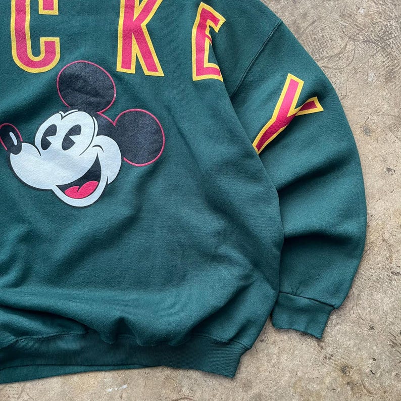 K&ouml;nnte beinhalten: Ein dunkelgr&uuml;nes Sweatshirt mit einer gro&szlig;en Mickey Mouse-Grafik. Das Wort "MICKEY" ist oben in gelben Buchstaben mit rosa Umrissen aufgedruckt. Der &Auml;rmel hat ein gelb-rosa Design.