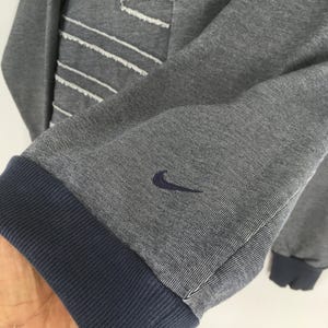 May include: Gray sweatshirt with a blue ribbed cuff and the Nike swoosh logo embroidered on the sleeve. The sweatshirt has white stitching details and a blue ribbed hem.
