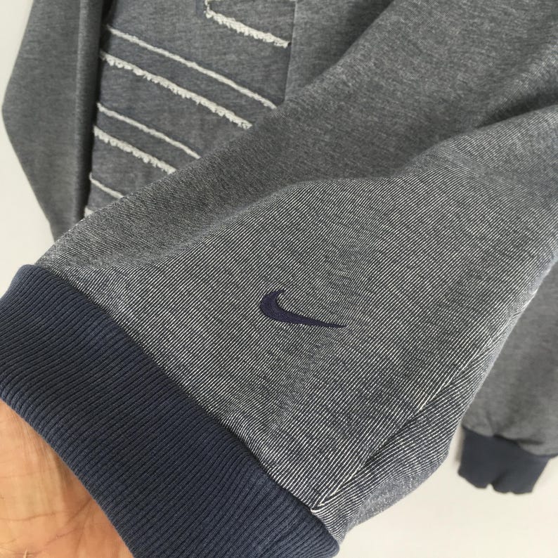May include: Gray sweatshirt with a blue ribbed cuff and the Nike swoosh logo embroidered on the sleeve. The sweatshirt has white stitching details and a blue ribbed hem.