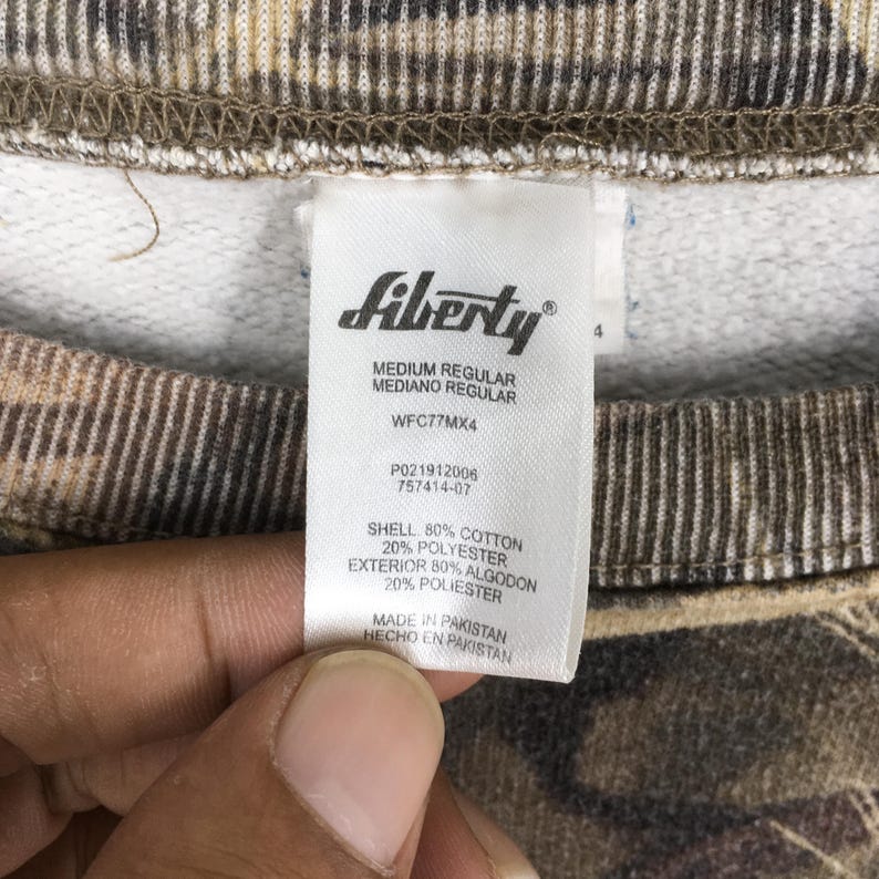 May include: Close-up of a tag from a Liberty sweatshirt. The tag is white with black text, including "MEDIUM REGULAR" and fabric composition details. The sweatshirt has a camouflage pattern in shades of brown and tan.