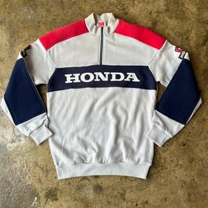 May include: A gray, red, and navy blue Honda sweatshirt with a quarter-zip collar. The word "HONDA" is prominently displayed across the chest in white block letters. The sleeves have navy blue patches and the shoulders are red.