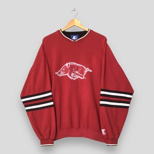 May include: A maroon long-sleeve crewneck sweatshirt featuring a white graphic of a charging razorback hog on the chest. The sleeves have black and white horizontal stripes near the cuffs, and the collar has a black and white trim detail.
