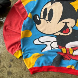 K&ouml;nnte beinhalten: Ein Vintage-Sweatshirt mit einer gro&szlig;en Mickey Mouse-Grafik. Das Sweatshirt hat einen blauen Hintergrund mit gelben Wolkenformen, rote &Auml;rmel und einen roten und gelben Besatz. Die Zeichentrickfigur l&auml;chelt.