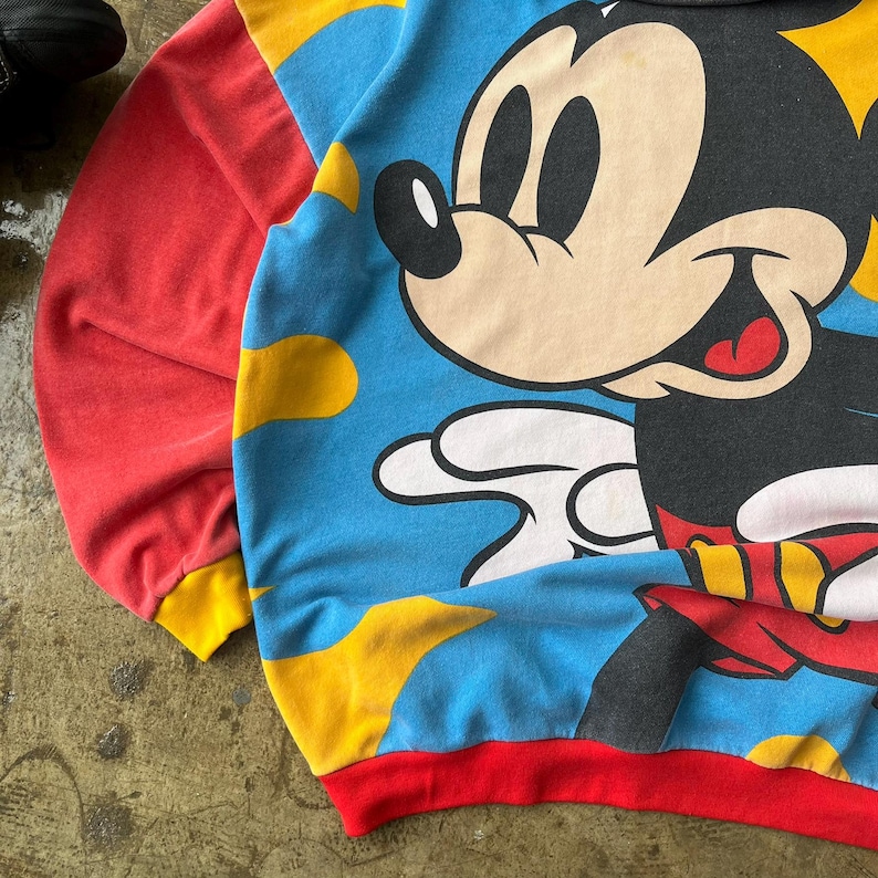 K&ouml;nnte beinhalten: Ein Vintage-Sweatshirt mit einer gro&szlig;en Mickey Mouse-Grafik. Das Sweatshirt hat einen blauen Hintergrund mit gelben Wolkenformen, rote &Auml;rmel und einen roten und gelben Besatz. Die Zeichentrickfigur l&auml;chelt.