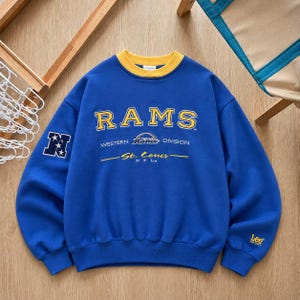 May include: A royal blue crewneck sweatshirt with a yellow collar. The front features yellow text reading RAMS, WESTERN DIVISION, and St. Louis. A navy blue patch with white stars and the letter N is on the right sleeve, and Lee Sport is on the left.